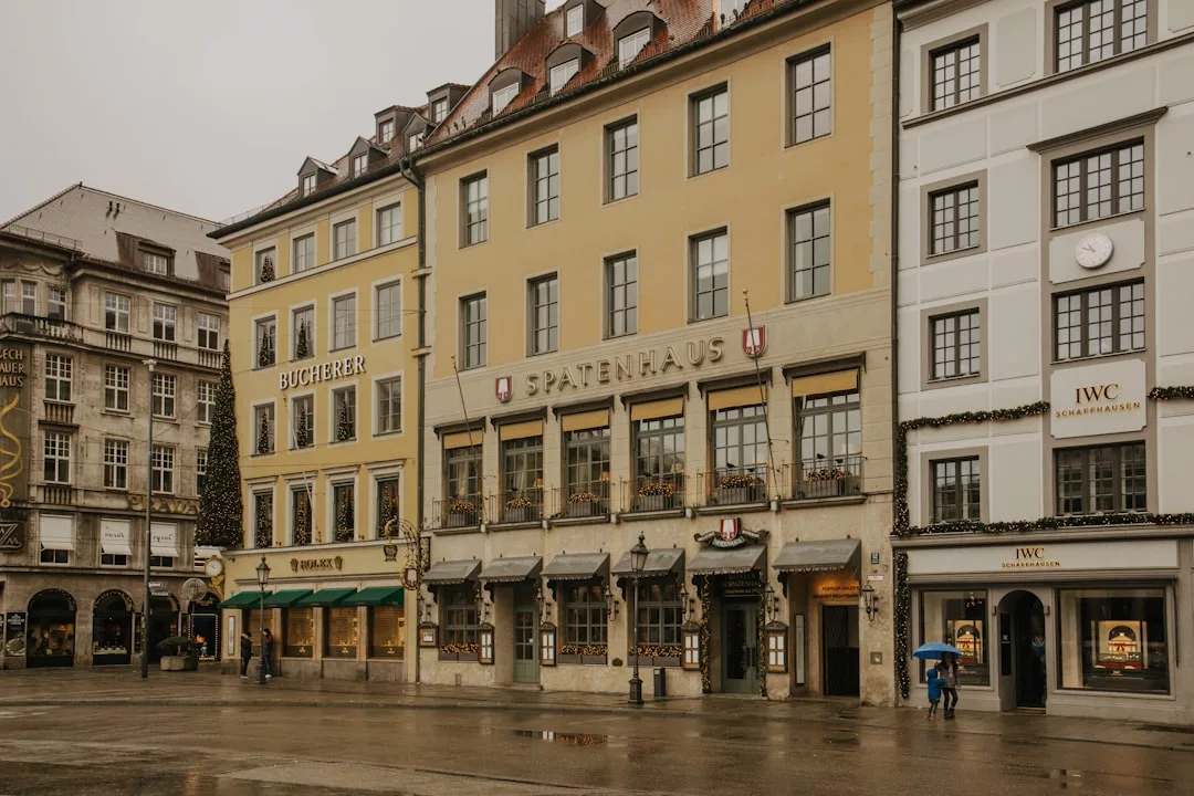 People with umbrellas walk past historic buildings and designer boutiques on a rainy day in a European city square, evoking the charm of upscale shopping in places like Puerto Portals.