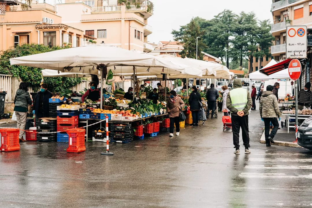 Outdoor market with people shopping at produce stalls under white tents on a rainy day in a city street, perfect for culinary tours or walking tours exploring the vibrant flavors of Palma Old Town.