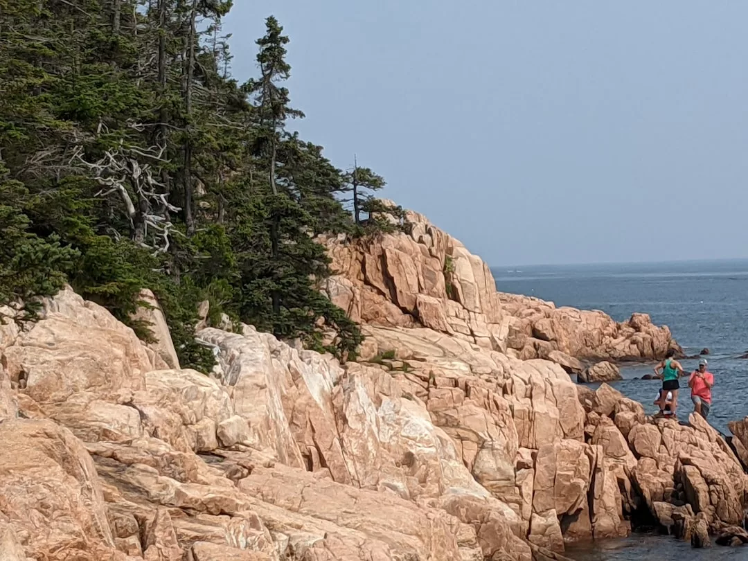 Two people stand on rocky, pinkish cliffs by the ocean at Cap de Formentor, with pine trees on the left and blue sky above—discovering one of Mallorca’s secret coastal trails.