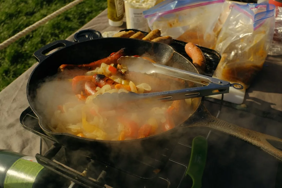 A smoky skillet of sizzling peppers, onions, and sausages is cooking outdoors near Playa de Muro, with the aroma rivaling gourmet paella from beachfront restaurants and food and bags close by.
