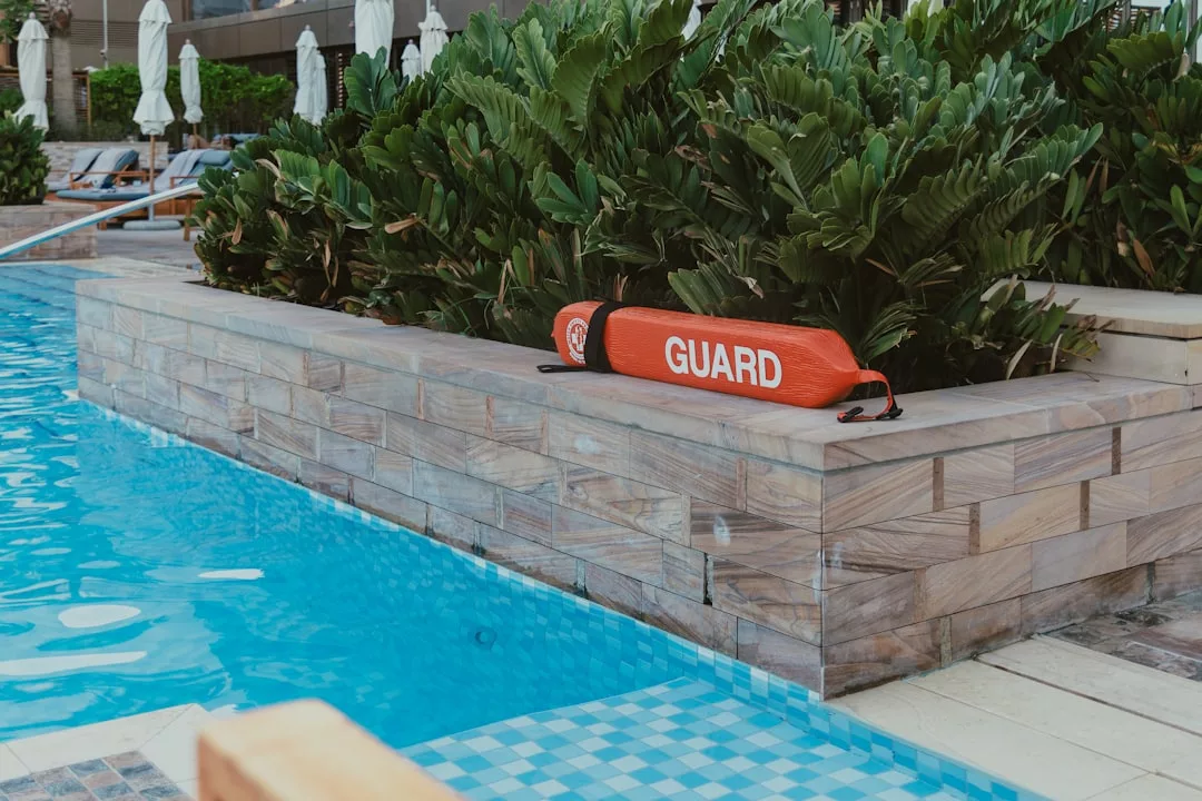A red lifeguard float rests by a swimming pool at a holiday resort, surrounded by green plants and stone walls.