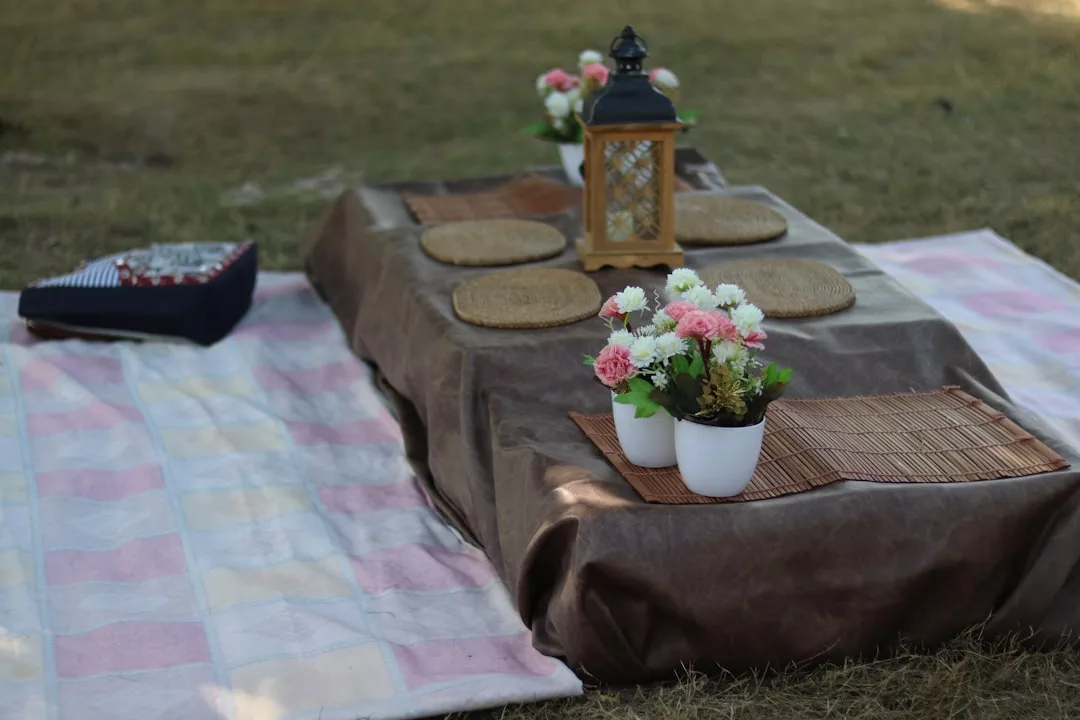 A charming beach picnic setup at Cala Llombards features mats, a brown-covered table, flowers, placemats, and a lantern arranged beautifully on the grass.