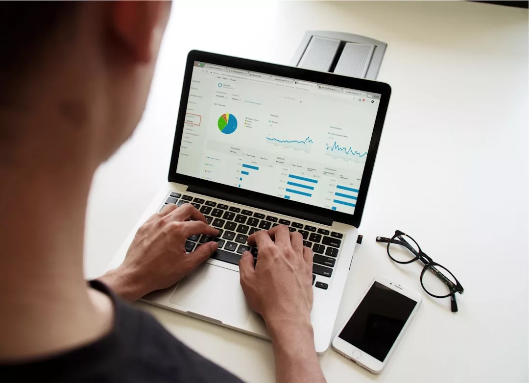 A person using a laptop displaying charts and graphs—perhaps analyzing commercial real estate trends or market entry strategies in Mallorca—with a smartphone and glasses on the desk nearby.