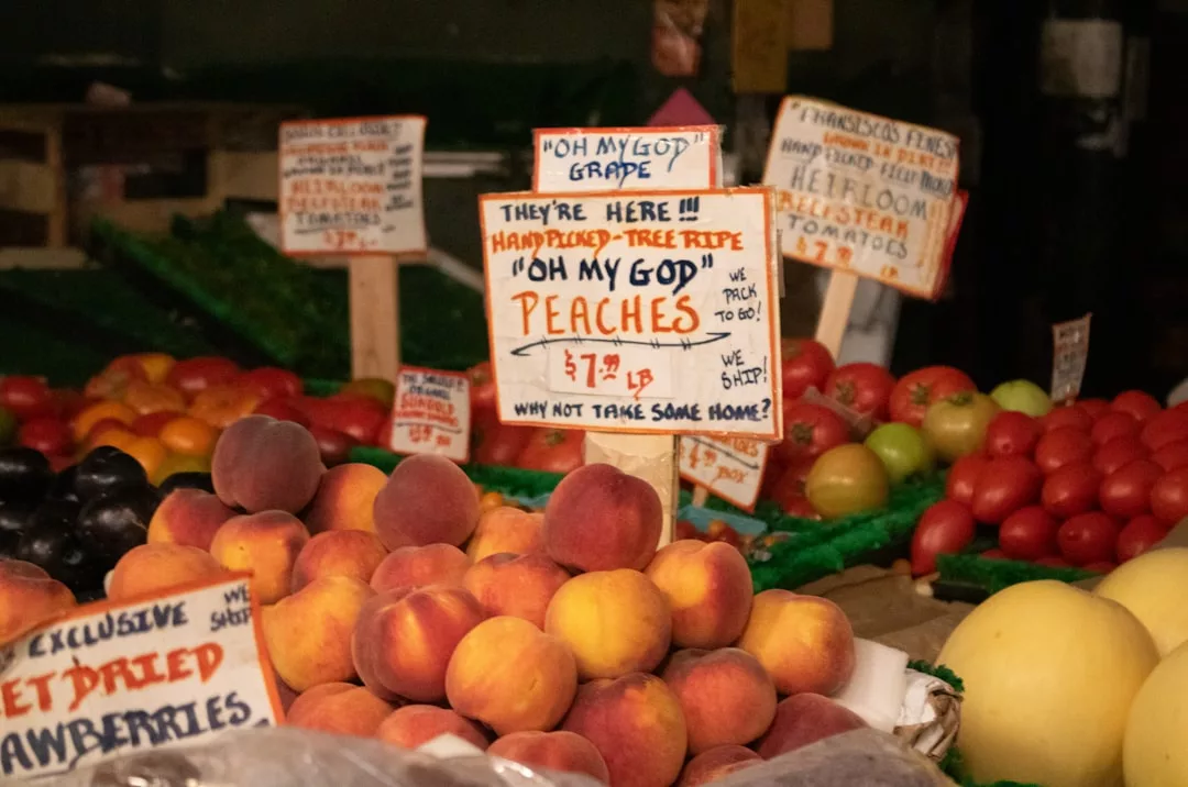 Display of peaches at a market with a hand-written sign reading, "Oh My God Peaches $7.99/lb," reminiscent of the fresh produce found in Sóller Valley or an organic vegan café.