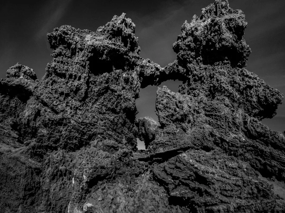 Jagged volcanic rock formations with a natural arch, captured in dramatic black and white—a striking reminder of the coastal properties at risk from sea-level rise.