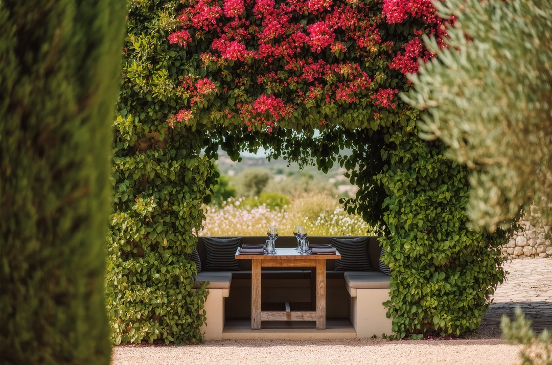 A wooden table and chairs set in a lush countryside garden, surrounded by red flowering plants and dense green foliage—perfect for enjoying the charm of Marratxí’s garden restaurants.